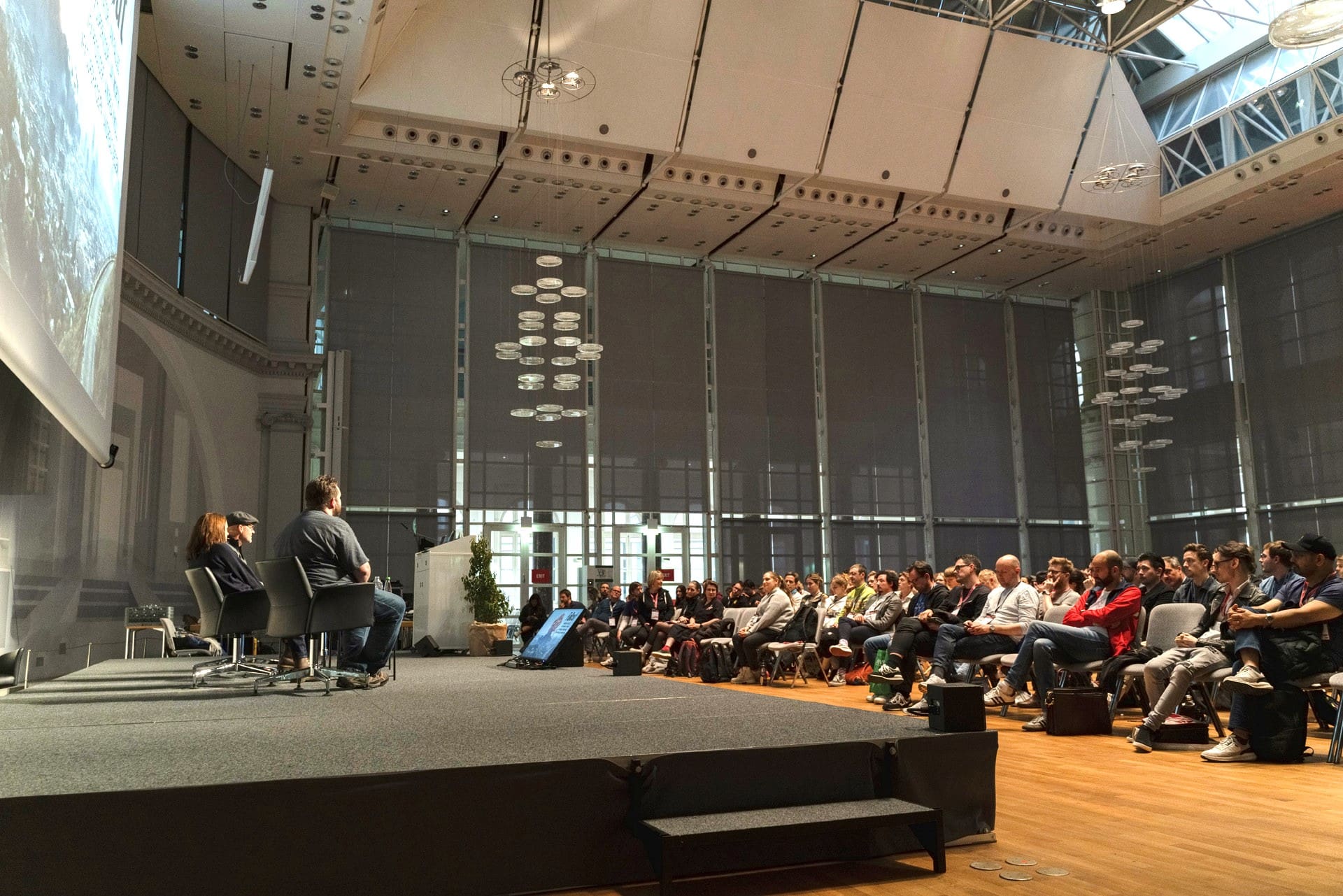 Blick in den großen Saal im Haus der Wirtschaft. Viele Menschen verfolgen einen Vortrag von drei Menschen auf einer Bühne.